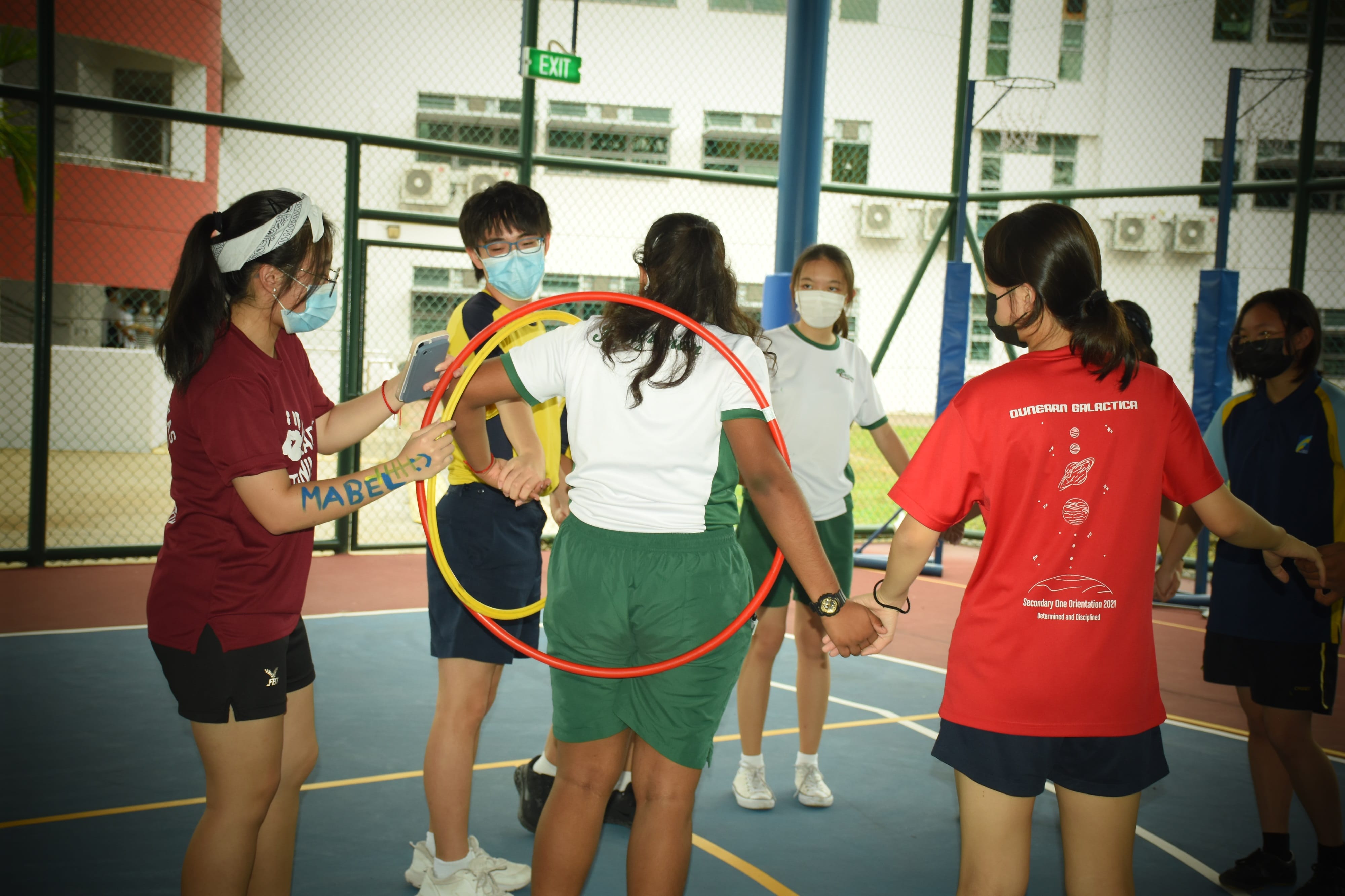 Students wearing masks play a hula hoop passing game on an outdoor school court during orientation.